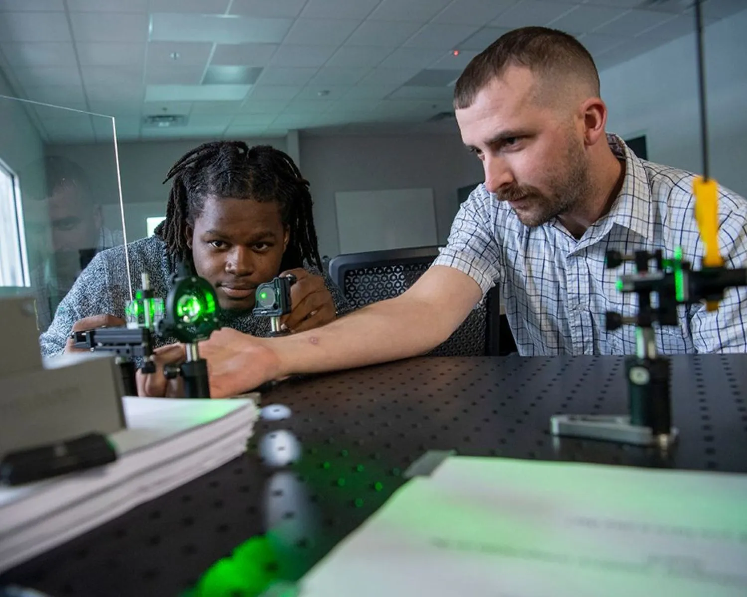 A student and Dr. Wei aligning optics on a laser table, a green beam visible through the mounted lenses and beam splitter.